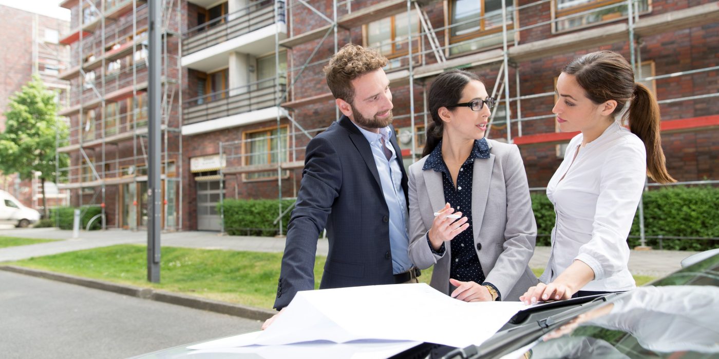 Three business people looking at a blueprint placed on a car bonnet, in front of a building which has a reddish brick façade and is covered in scaffolding, Fast Forward Commercial Excellence