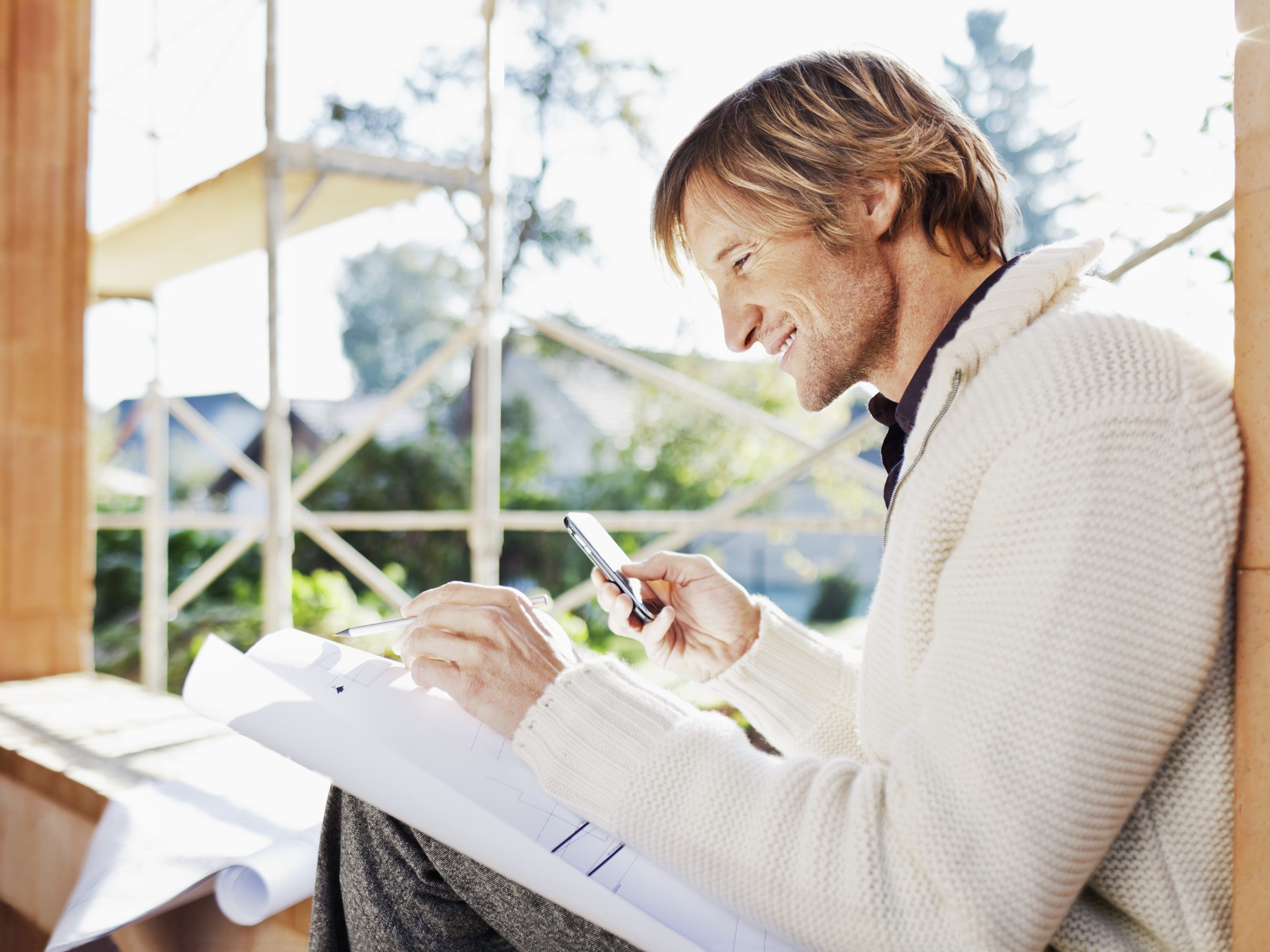 Smiling man with blueprint and mobile phone sitting on window sill at construction site